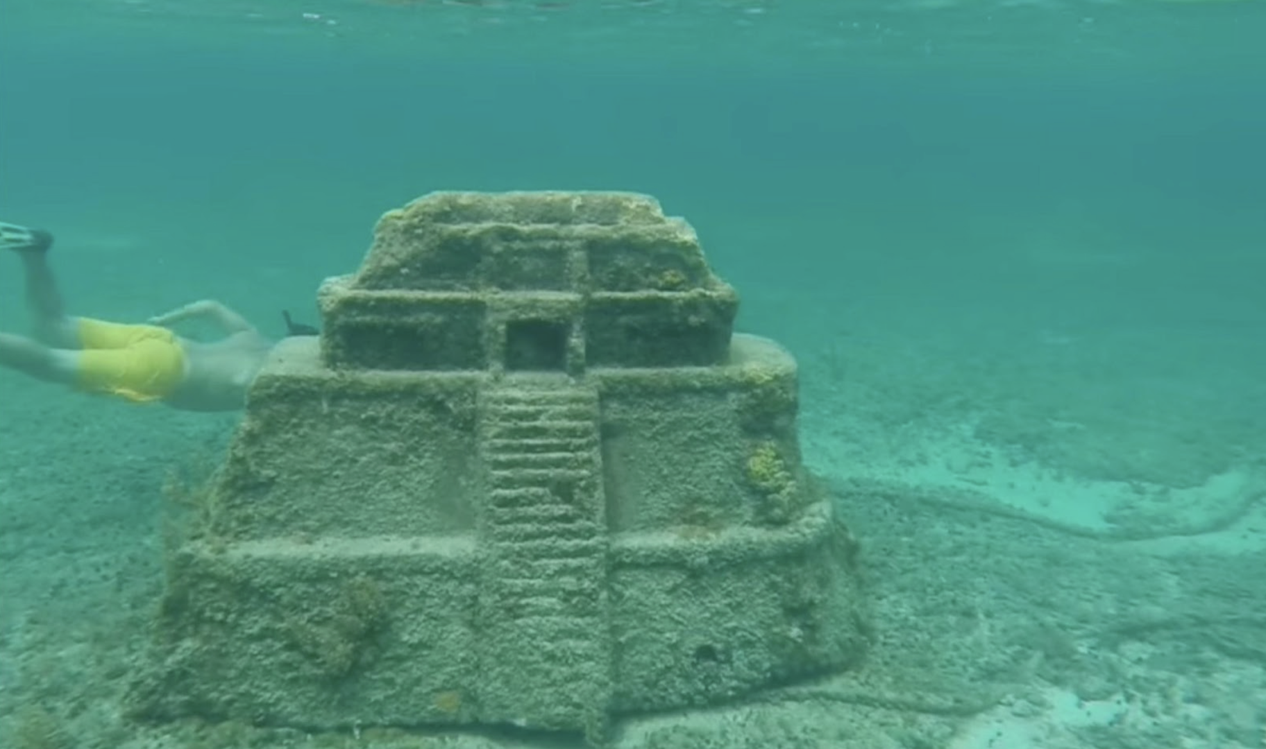 A photograph taken underwater showing a diver's arm reaching out towards a small, pyramid-like stone structure covered in marine growth. The pyramid, with a central doorway and steps leading up to it, sits on the ocean floor